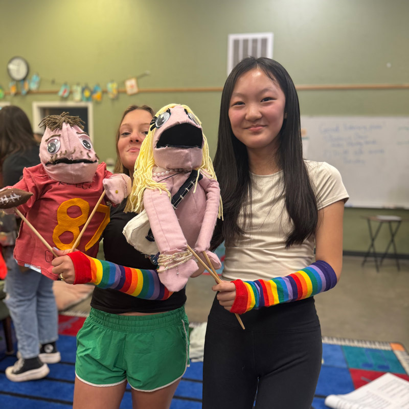 Students from Community School create a variety of different artworks: shown here is puppetry with two students. One student is caucasian with a pink puppet wearing a red shirt. Another student is Asian American and is holding her puppet with blond hair. Both girls are wearing rainbow gloves.