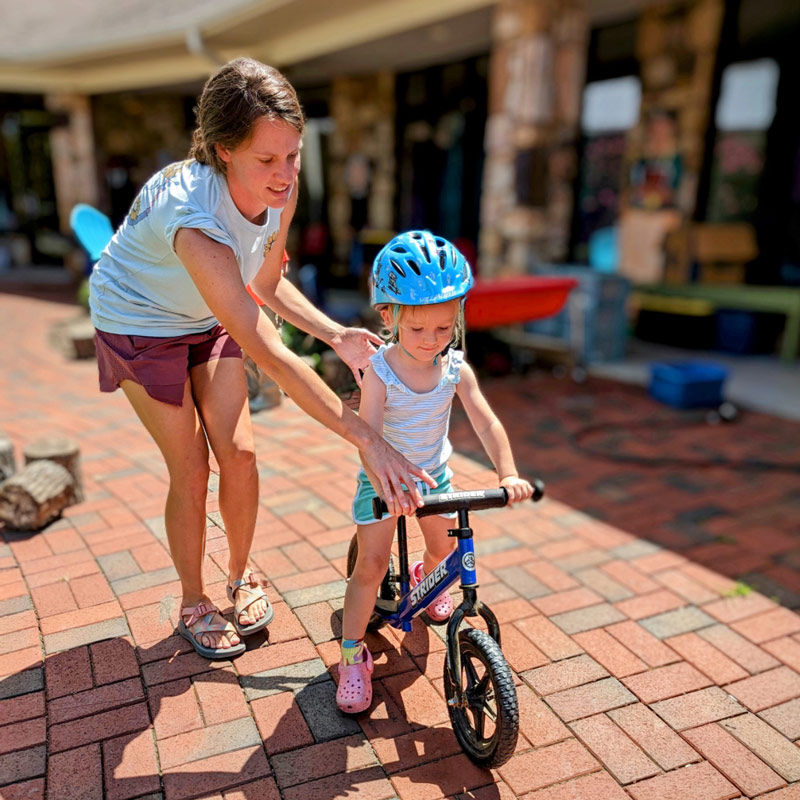 A caucasian child on a bike with training wheels with her teacher gently guiding her at Community School near Roanoke, Virginia.
